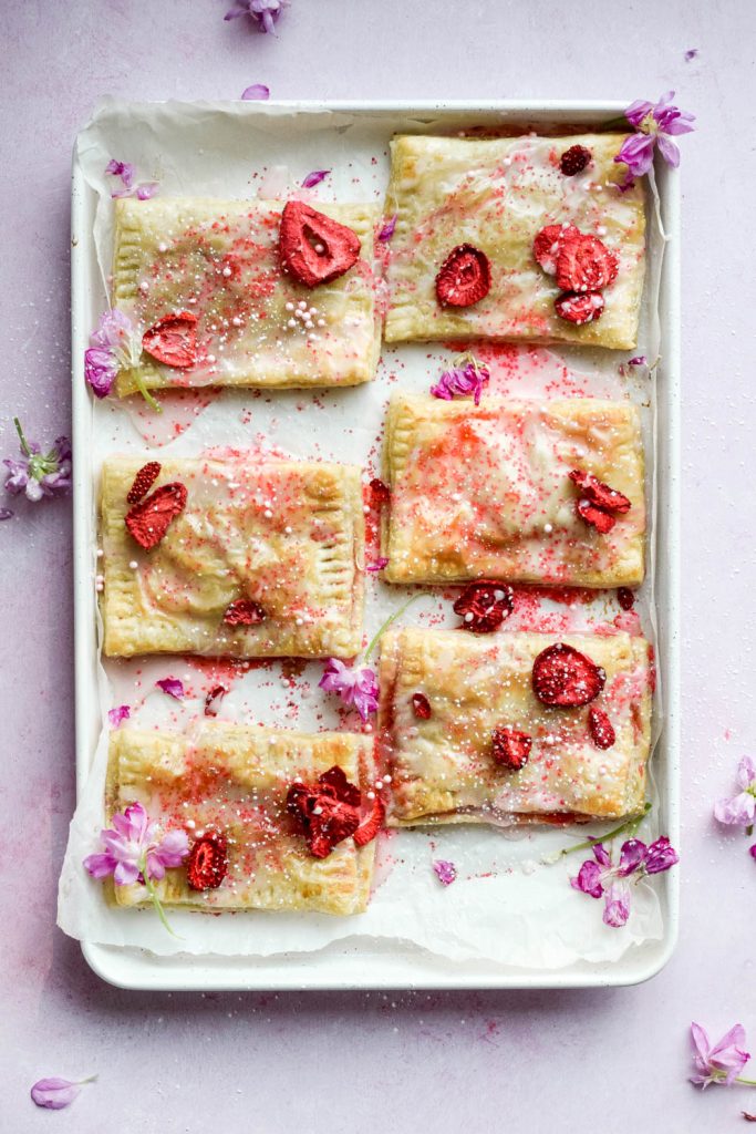Baked frosted strawberry pop tarts topped with edible flowers and dried strawberries on parchment on white baking tray.