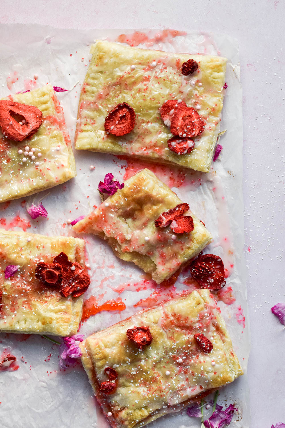 Baked frosted strawberry pop tarts topped with edible flowers and dried strawberries on parchment.