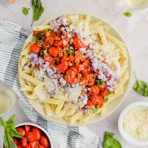 penne pasta topped with bruschetta ingredients before tossing to make pasta salad with additional recipe ingredients surrounding bowl on white background.