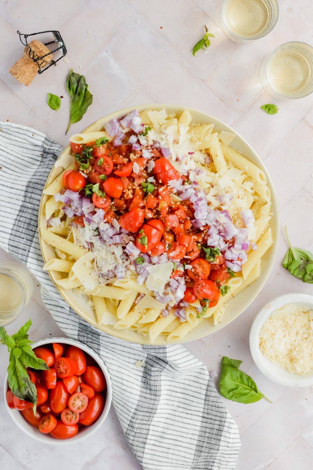 penne pasta topped with bruschetta ingredients before tossing to make pasta salad with additional recipe ingredients surrounding bowl on white background.