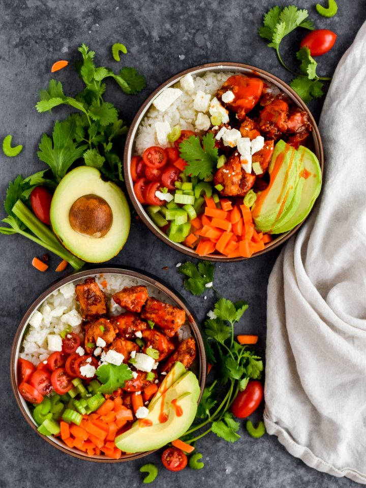 two buffalo chicken rice bowls garnished with feta and fresh veggies surrounded by herbs on slate gray background.