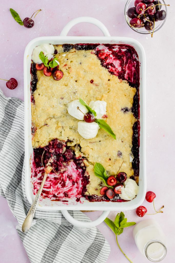white baking dish filled cherry blueberry cobbler topped with scoops of vanilla ice cream with a spoon inserted into corner of dish with a few bites taken out.