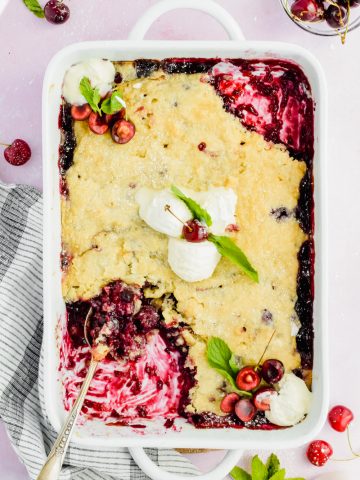 white baking dish filled cherry blueberry cobbler with a spoon inserted into corner of dish with a few bites taken out.