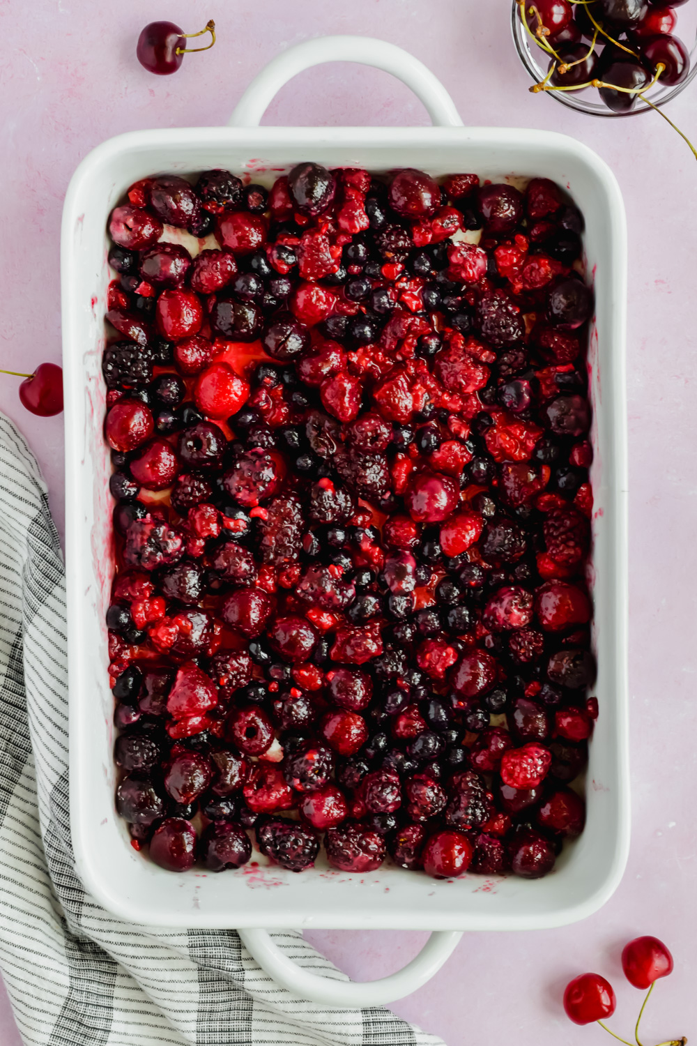 white baking dish filled with mixed berries and cherries surrounded by additional cherries.