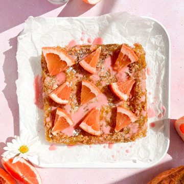 baked grapefruit bars on white parchment topped with grapefruit slices and pink glaze beside grapefruit slices on light pink background.