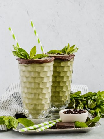 two green healthy mint chocolate smoothies in tall glasses rimmed with chocolate and garnished with fresh mint leaves and green and white striped straws on silver tray.