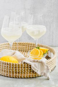 three wine glasses half full of ice and limoncello spritz in wicker basket with lemon slices in basket.