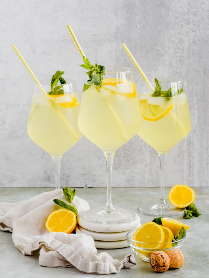 three full limoncello spritz drinks in wine glasses garnished with lemon wedges, mint leaves, and straws.