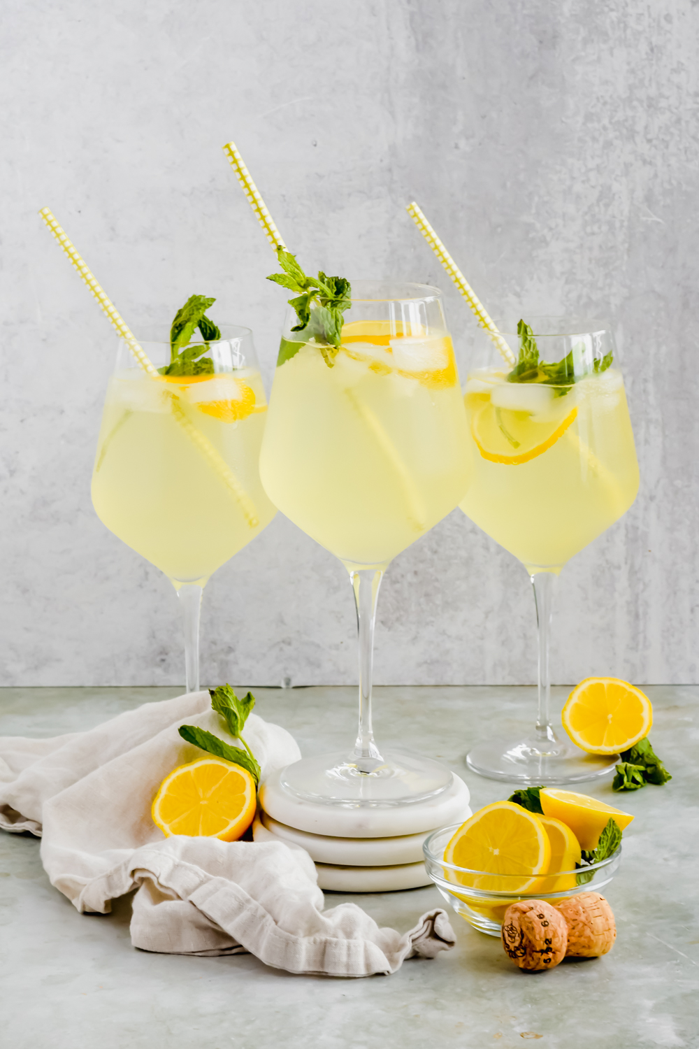 three full limoncello spritz drinks in wine glasses garnished with lemon wedges, mint leaves, and straws with bottle additional lemon wedges surrounding.