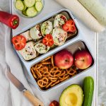 mediterranean pinwheels in silver lunch box compartment with pretzels and apples in other lunch box compartments surrounded by additional recipe ingredients.