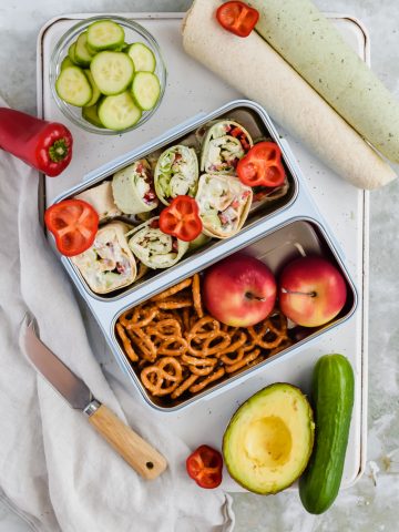 mediterranean pinwheels in silver lunch box compartment with pretzels and apples in other lunch box compartments surrounded by additional recipe ingredients.
