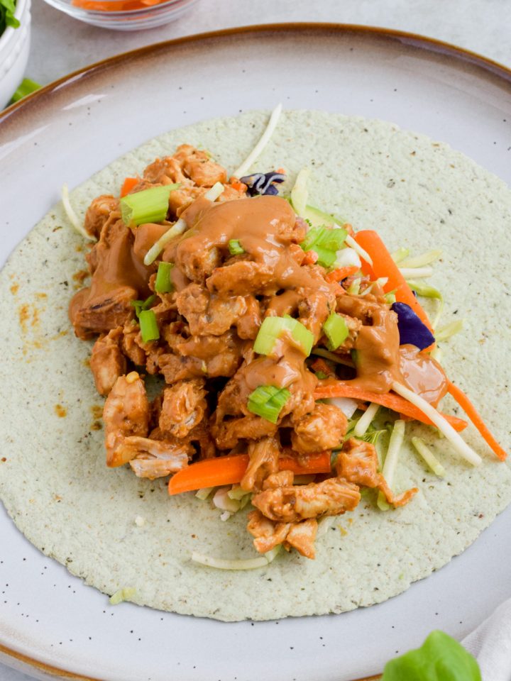 filled oriental chicken tortilla wrap drizzled with peanut sauce before wrapping on white plate.