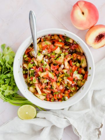 jalapeno peach salsa tossed in large white mixing bowl with silver spoon in bowl beside fresh cilantro leaves, half a lime and a whole peach.
