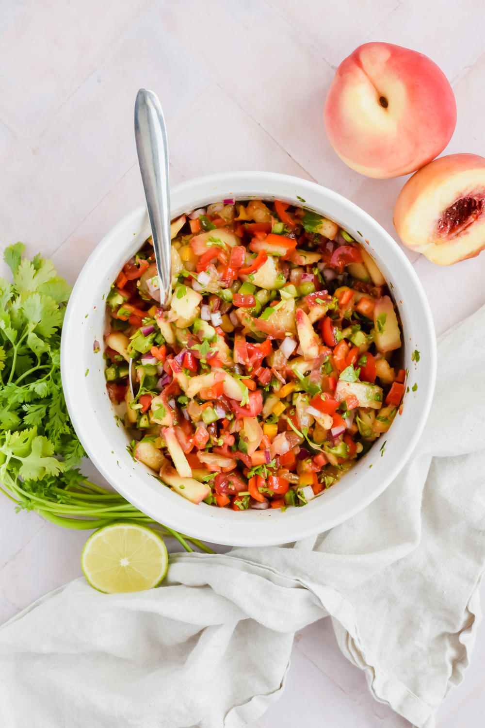 jalapeno peach salsa tossed in large white mixing bowl with silver spoon in bowl beside fresh cilantro leaves, half a lime and a whole peach.