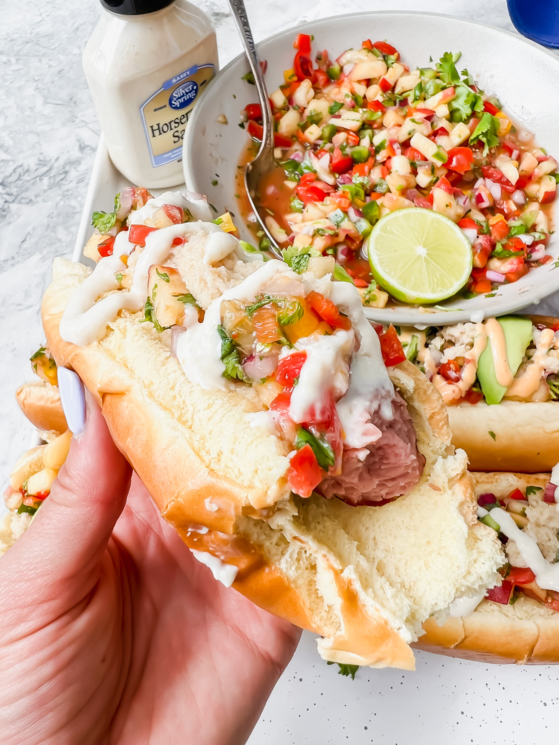 close up shot of hot dog in white bun garnished with jalapeno peach salsa and horseradish with a bite taken out with big white bowl of additional salsa in background.