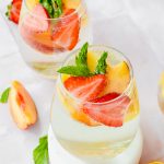 two stemless wine glasses full of sparkling white wine peach sangria garnished with strawberry and peach slices and mint leaves.