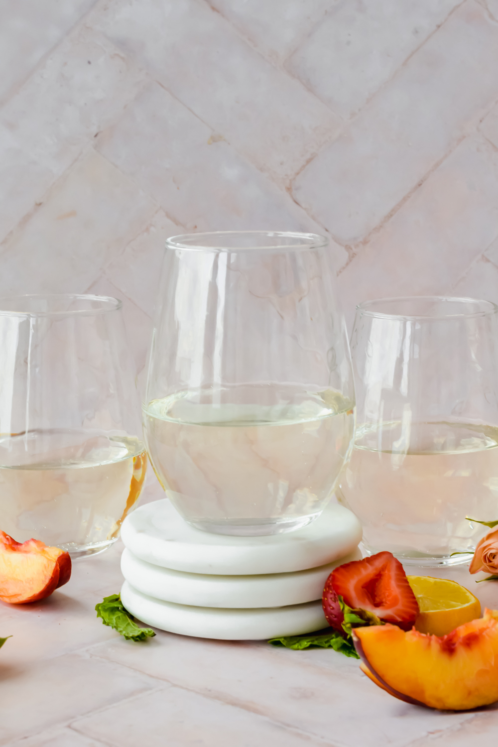 three wine glasses half full of white wine with strawberry and peach slices surrounding on stone white background.