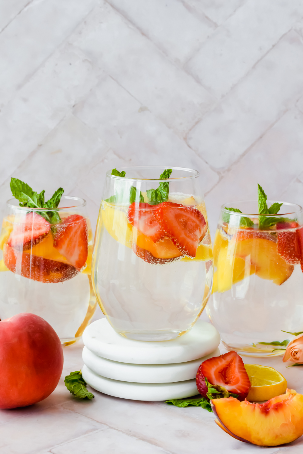 three stemless wine glass full of sparkling white wine peach sangria garnished with strawberry and peach slices and mint leaves with middle glass on three stacked white coasters.
