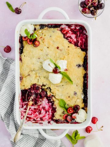 white baking dish filled cherry blueberry cobbler topped with scoops of vanilla ice cream with a spoon inserted into corner of dish with a few bites taken out.