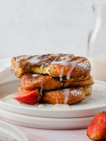 stack of three slices of churro french toast with a bite out of the top slice garnished with white glaze, powdered sugar and strawberry slices with glass of milk in background.