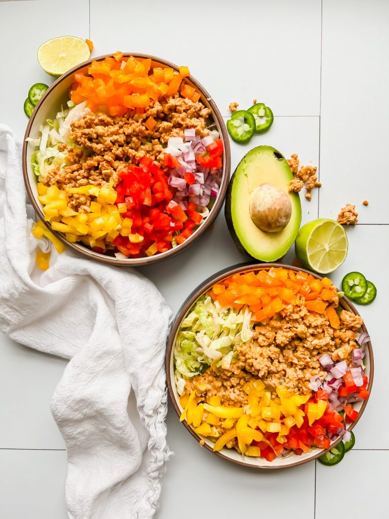 two bowls of taco salad filled with lettuce, turkey, and fresh peppers on a white tiled background