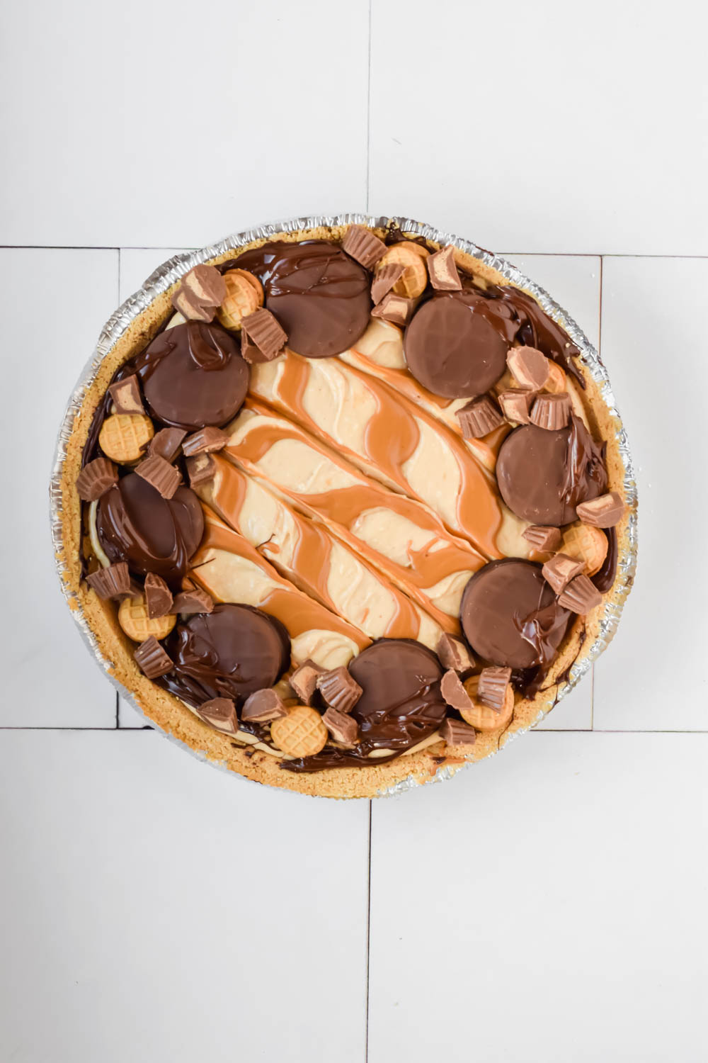 nutter butter pie swirled with peanut butter on top with additional chocolate peanut butter candies garnishing pie in foil pie pan on white background.
