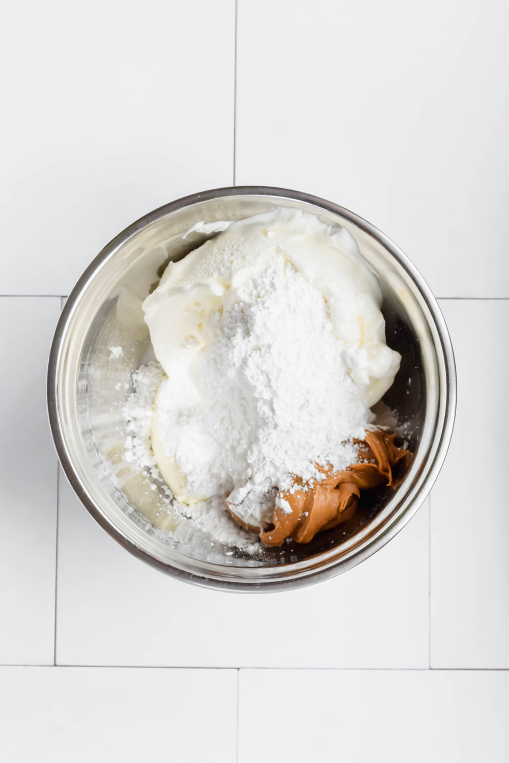 peanut butter, powdered sugar, and cream cheese block and yogurt in silver mixing bowl on white background.