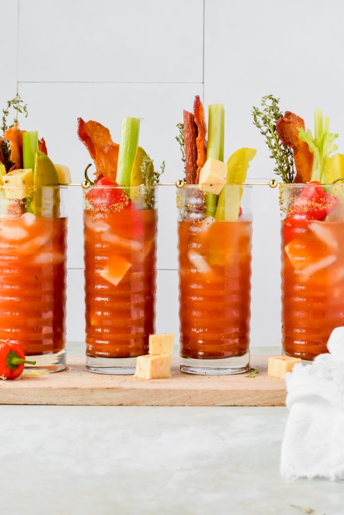 four pickle bloody mary aligned on wood plank garnished with celery sticks, pickle spears, bacon, cheese cubes, and mini bell peppers.