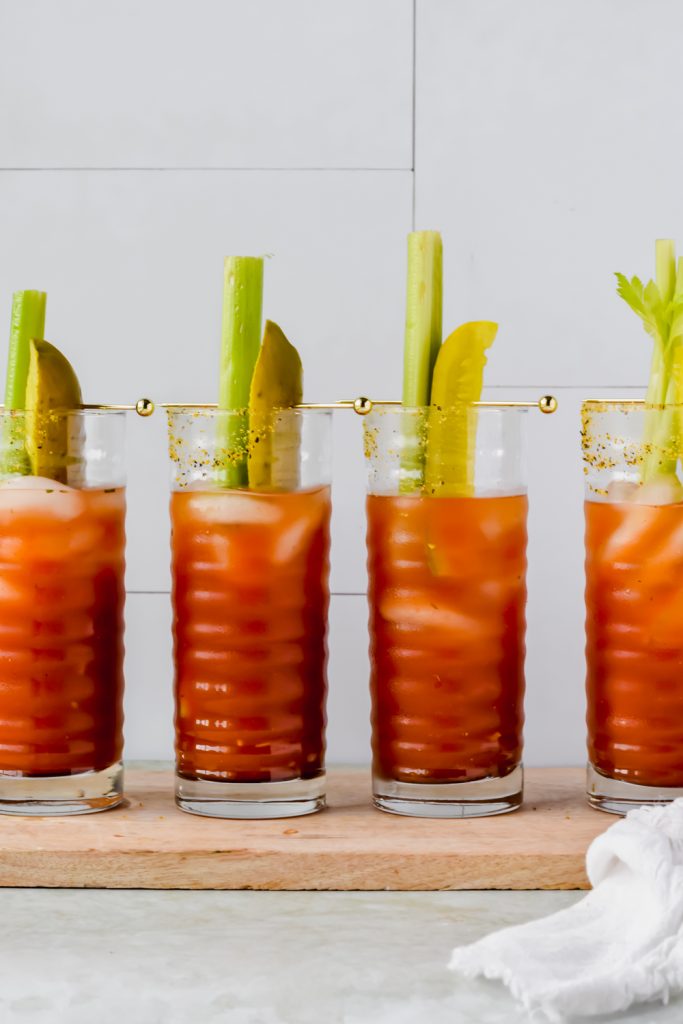 four pickle bloody mary aligned on wood plank garnished with celery sticks and pickle spears.