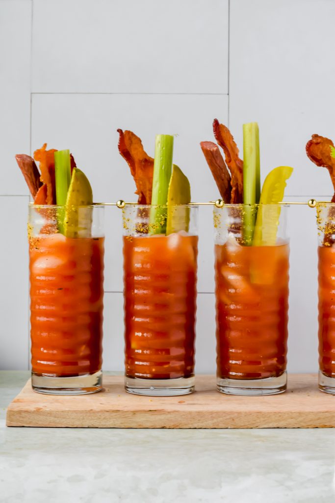 three pickle bloody marys aligned on wood plank garnished with celery sticks, pickles, and bacon.