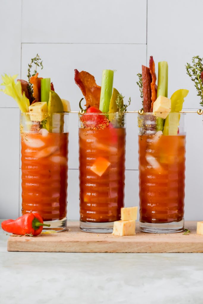 three pickle bloody mary aligned on wood plank garnished with deli toppings.