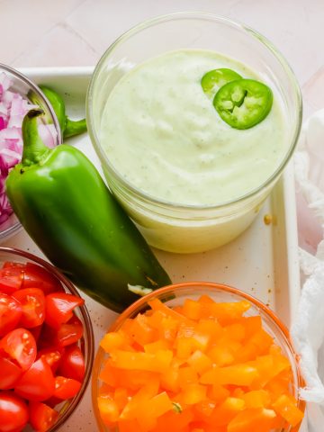 creamy jalapeno sauce in glass beaker beside fresh jalapeno and small bowls of chopped peppers, tomatoes, and onion.