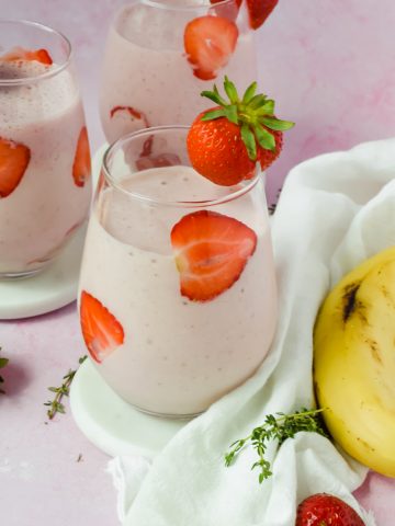 three glasses filled with strawberry banana protein smoothie with strawberry slices lining the glasses and a strawberry wedged onto edge of glass beside whole banana and white linen towel.