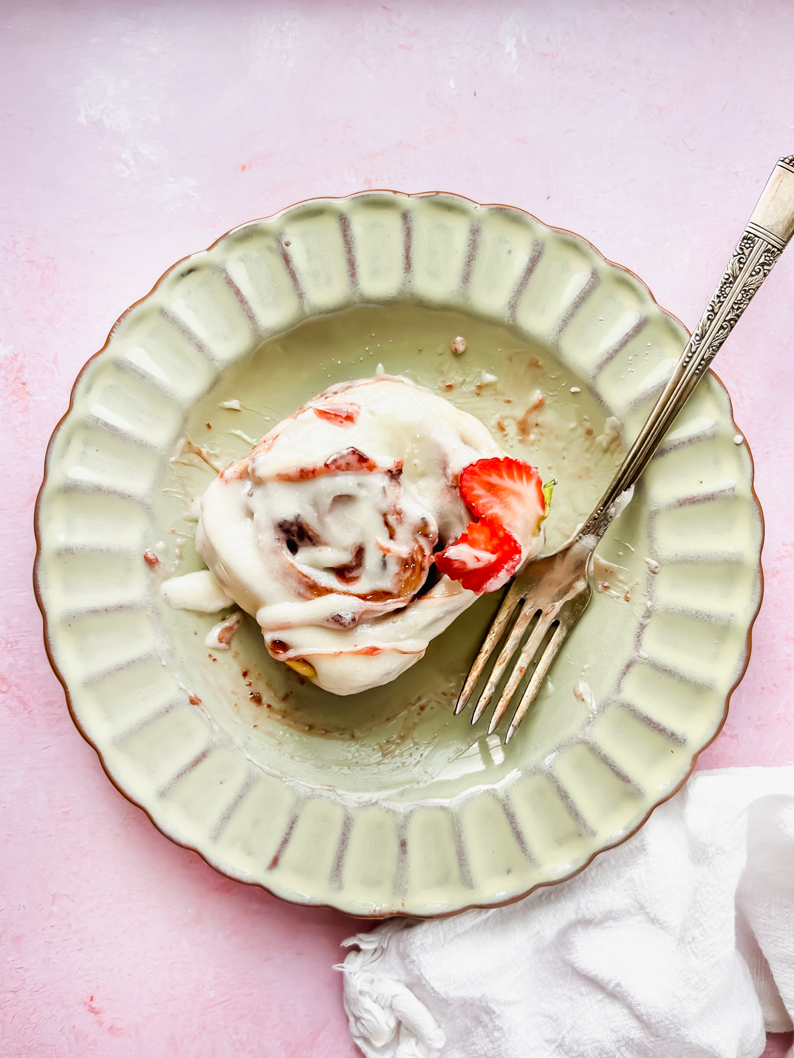 One glazed strawberry cinnamon roll on antique plate garnished with fresh strawberry slices.