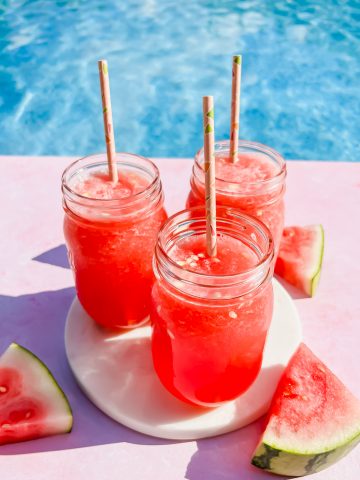 three watermelon vodka slushies in mason jars on white coaster beside slices of watermelon with pool in background.