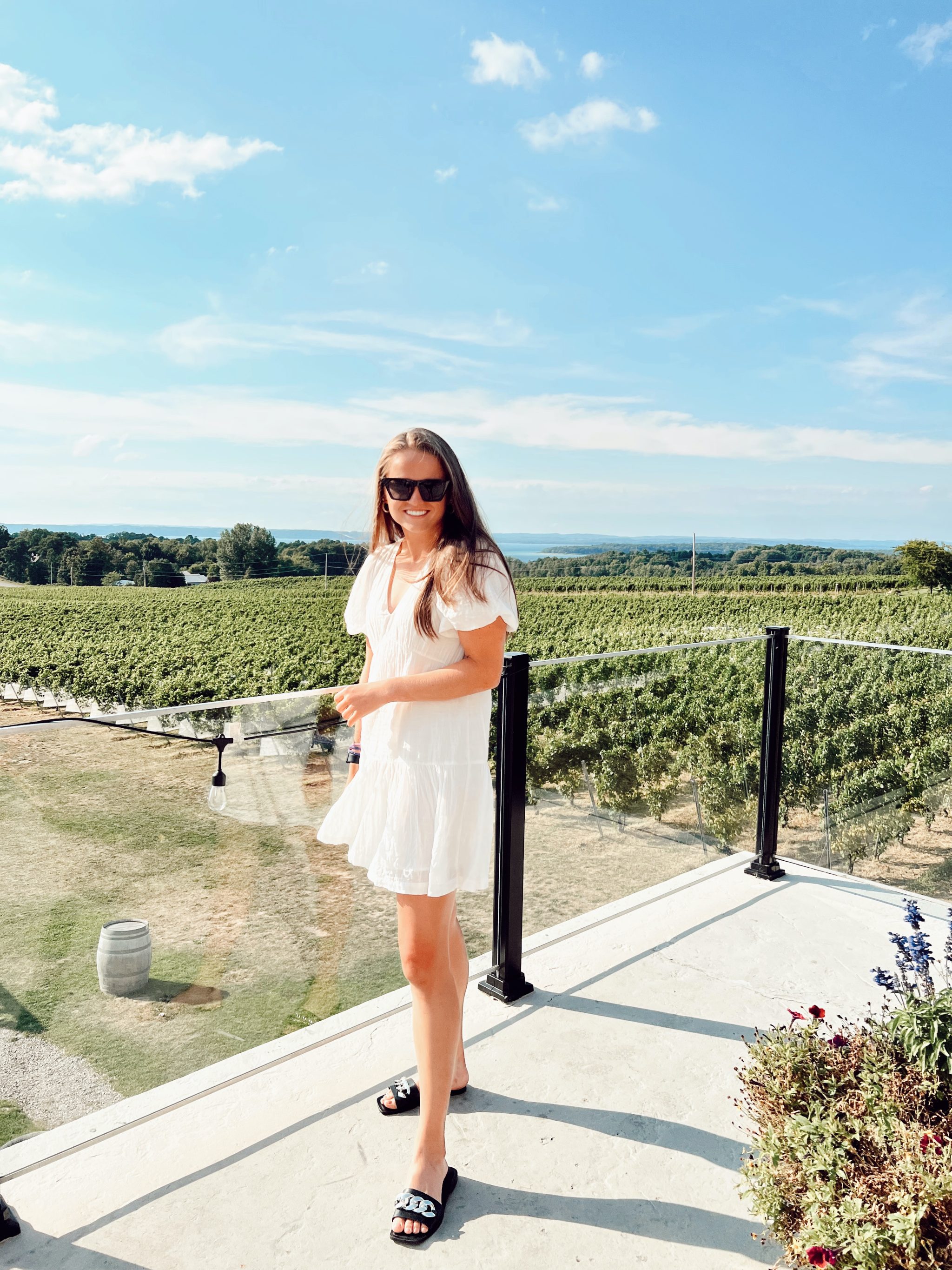 girl in white dress in front of glass rail with rows of wine grape vines in background.