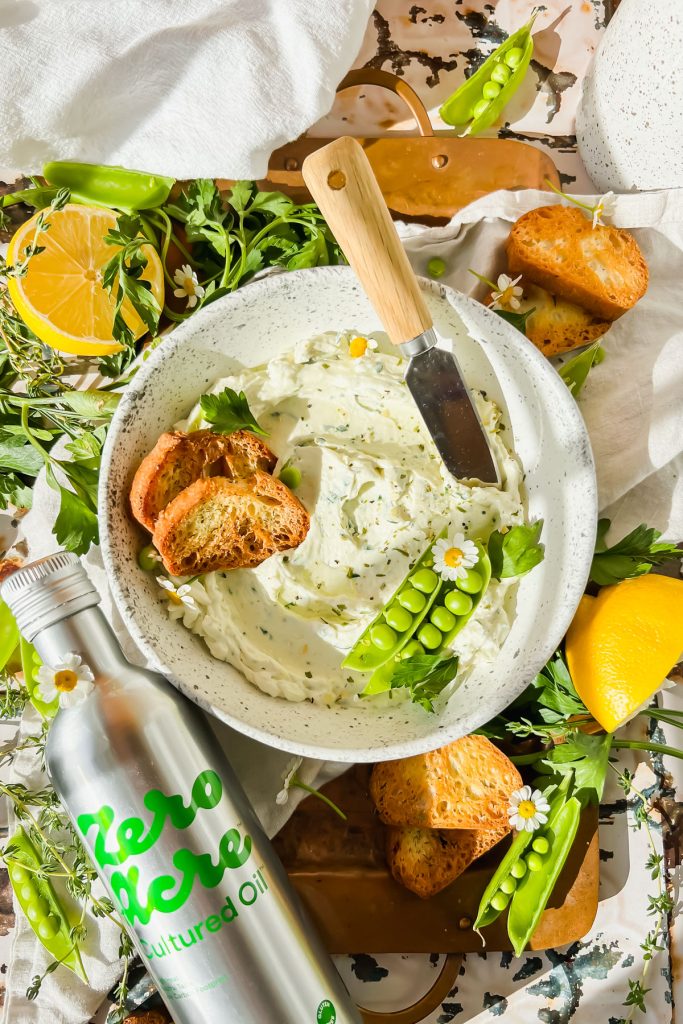 cream cheese garlic dip in white bowl with two toasted crostinis and pea pods in bowl beside bottle of zero acre cultured oil with fresh herbs surrounding.