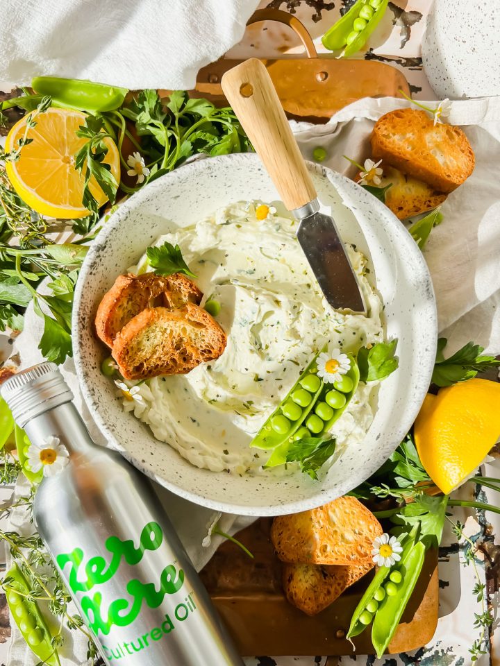 cream cheese garlic dip in white bowl with two toasted crostinis and pea pods in bowl beside bottle of zero acre cultured oil with fresh herbs surrounding.