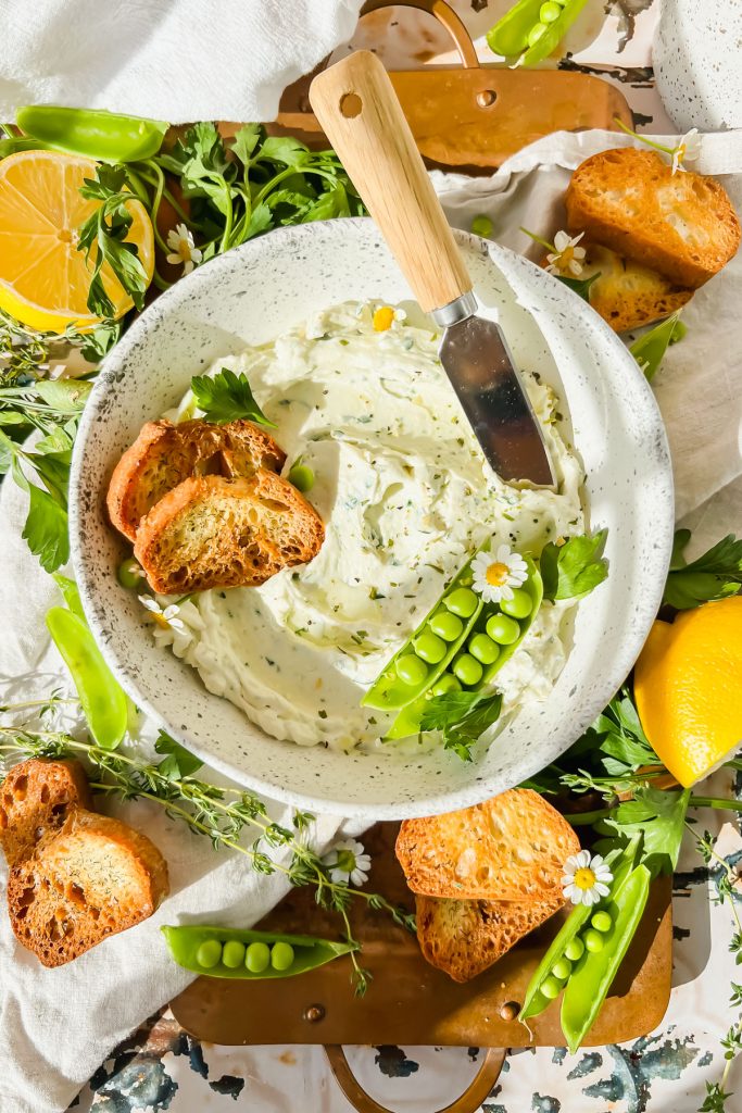 cream cheese garlic dip in white bowl with butter knife stuck into bowl surrounded by lemon wedges, pea pods, bread slices, and fresh herbs.