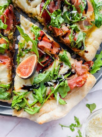 slice taken out of whole fig prosciutto pizza topped with arugula and balsamic drizzle.