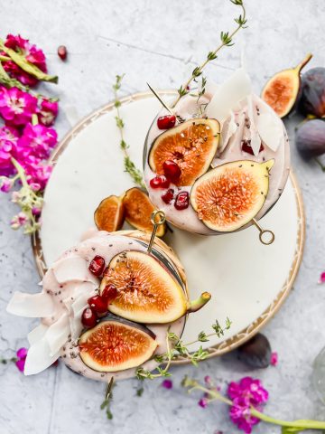 overhead shot of two fig smoothies garnished with fresh fig halves, pomegranate seeds, and coconut flakes on white serving tray with flowers and greenery surrounding.