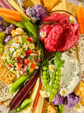 roasted beet dip, cream cheese garlic dip, and avocado crack dip in glass bowls surrounded by fresh veggies and crackers for dipping.