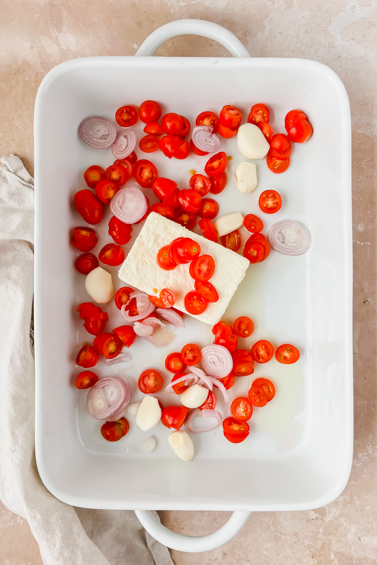 block of feta, garlic bulbs, sliced shallots, and cherry tomato halves in white baking dish.