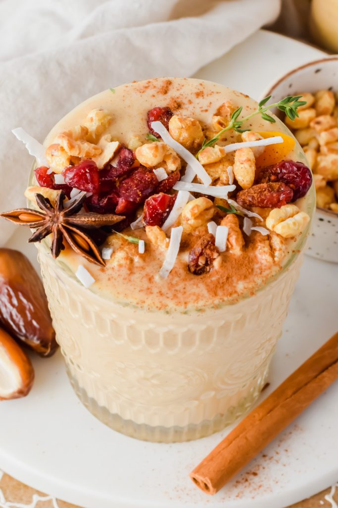 butternut squash smoothie garnished with nuts, dried fruit, coconut flakes, and star anise beside whole cinnamon stick, sliced open date and small bowl of nuts in background.