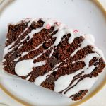slice of chocolate peppermint bread with icing drizzled across and crushed candy canes on top on white serving plate.