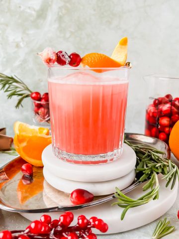 cranberry whiskey sour garnished with orange peel and cranberries on white coaster on silver tray surrounded by additional cocktail garnishes.