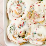 close up of baked funfetti cinnamon rolls in white baking dish smothered with white icing and sprinkles.
