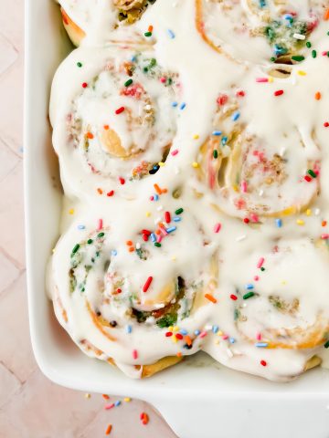 close up of baked funfetti cinnamon rolls in white baking dish smothered with white icing and sprinkles.