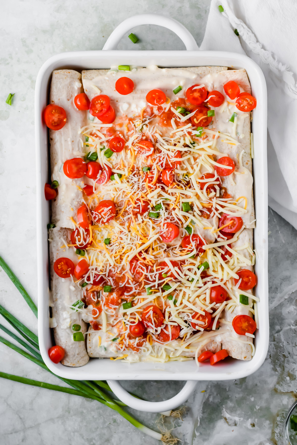 healthy ground turkey enchiladas rolled in white baking dish topped with extra sauce, cheese, and cherry tomato halves prior to baking.