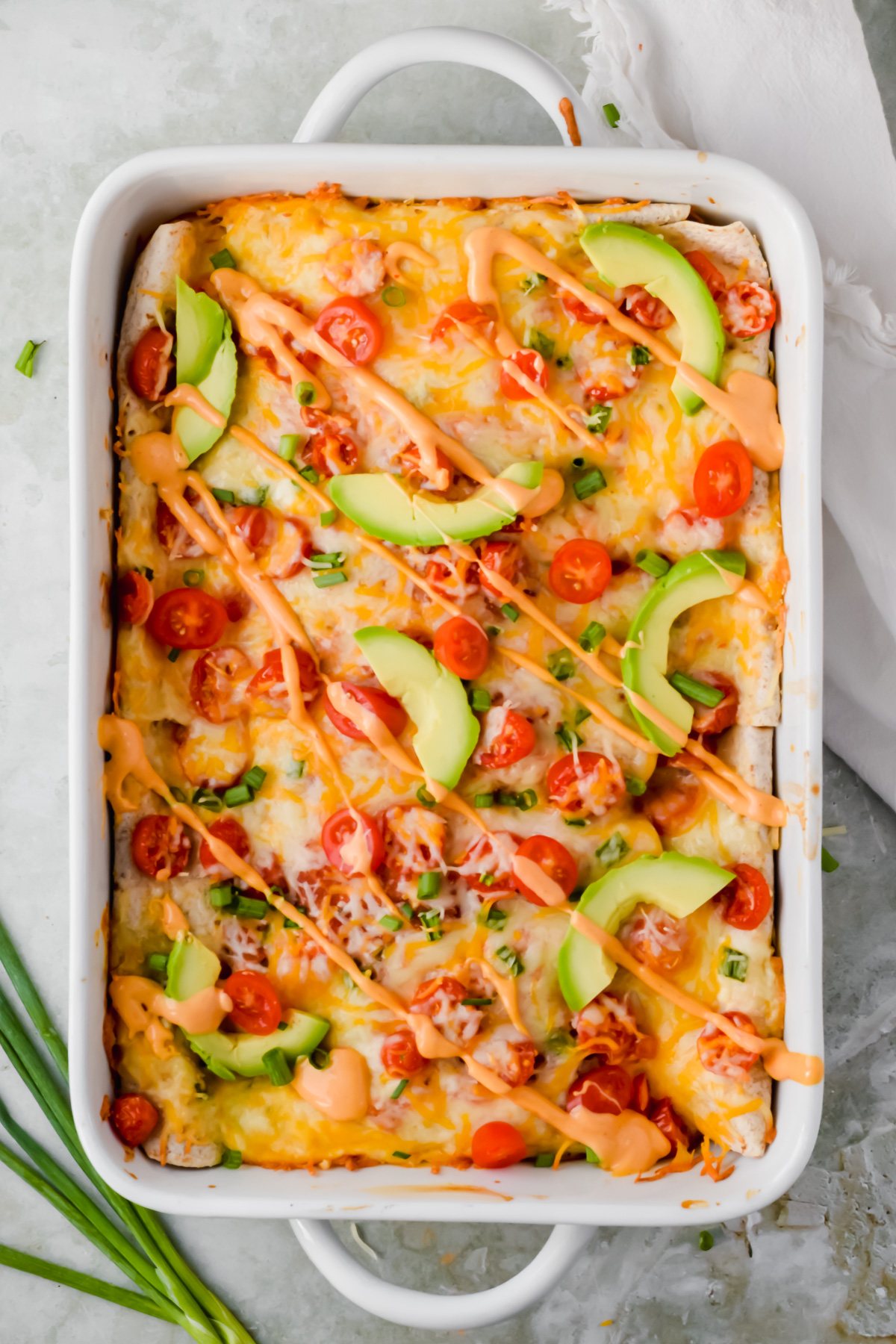 baked healthy ground turkey enchiladas garnished with cherry tomato slices, avocado slices, green onion and queso in white baking pan.